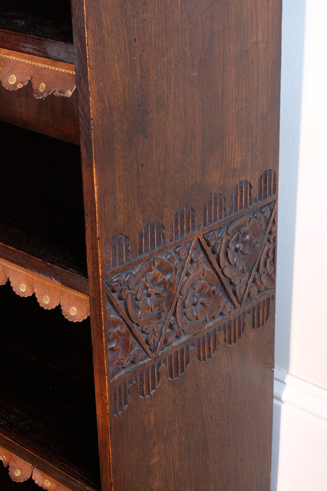 lower chip carved view of a Edwardian Arts and Crafts carved oak bookcase with original tooled leather dust fringing shelves