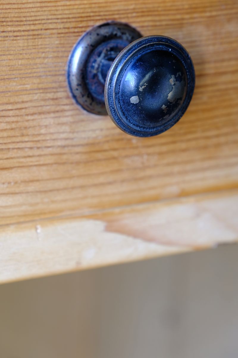 A close-up of a round, dark distressed cabinet knob mounted on a light wood drawer front. Late Victorian pine marble top bedside cupboard with Carrara marble and concealed brass pivot hinges