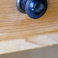 A close-up of a round, dark distressed cabinet knob mounted on a light wood drawer front. Late Victorian pine marble top bedside cupboard with Carrara marble and concealed brass pivot hinges