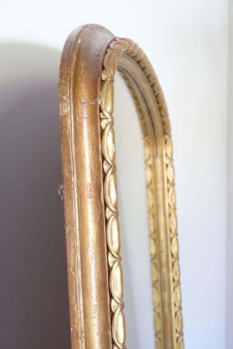 The frame itself has a substantial architectural section, built around a broad convex cushion-moulded outer profile. Set immediately inside this is the continuous carved guilloche-derived border of elongated oval reserves, followed by a restrained inner moulding framing the original arched plate. Seen from the side, the depth of these successive mouldings becomes particularly apparent, giving the mirror considerably more sculptural presence than its relatively restrained frontal outline initially suggests.