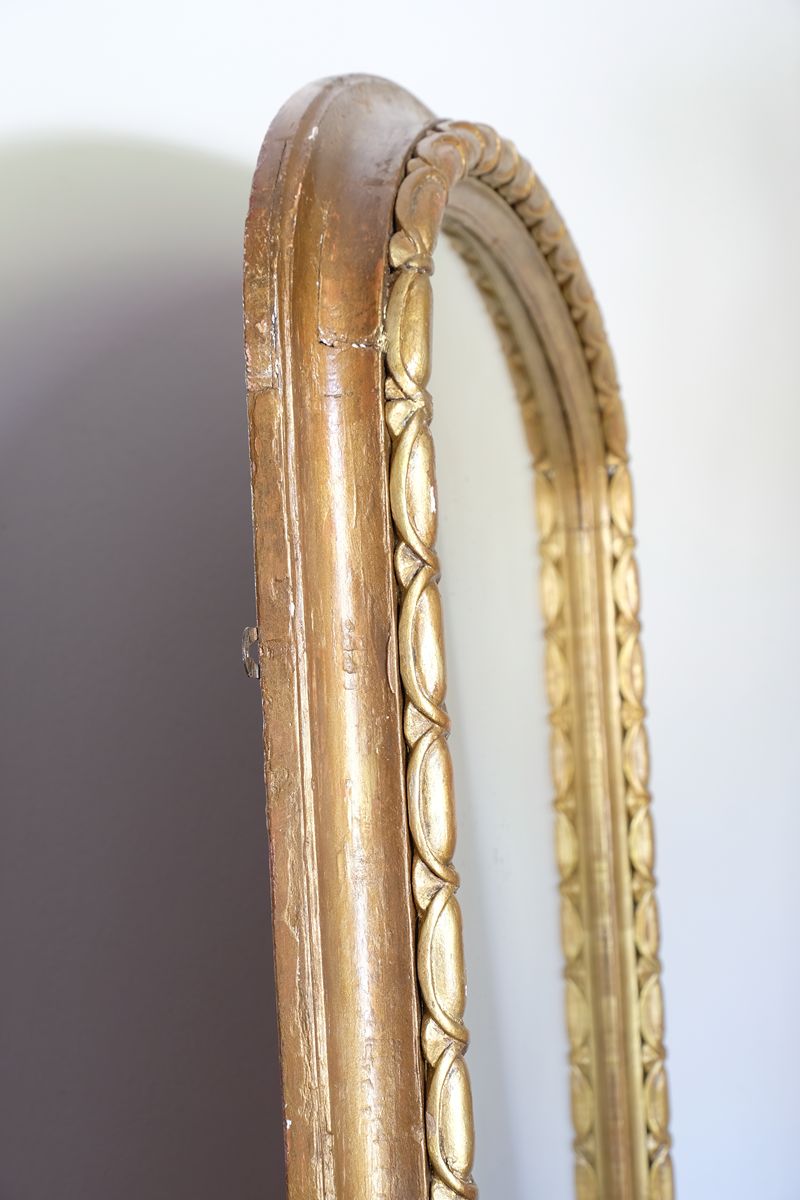 The frame itself has a substantial architectural section, built around a broad convex cushion-moulded outer profile. Set immediately inside this is the continuous carved guilloche-derived border of elongated oval reserves, followed by a restrained inner moulding framing the original arched plate. Seen from the side, the depth of these successive mouldings becomes particularly apparent, giving the mirror considerably more sculptural presence than its relatively restrained frontal outline initially suggests.